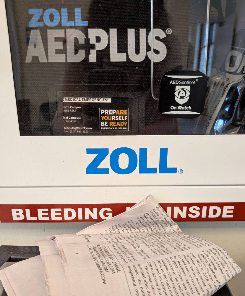 A sign on the door to an emergency first aid panel that is positioned just above a trash can in such a way that a piece of trash is blocking one of the words, so it looks like the sign says "bleeding inside" instead of "bleeding kit inside."
