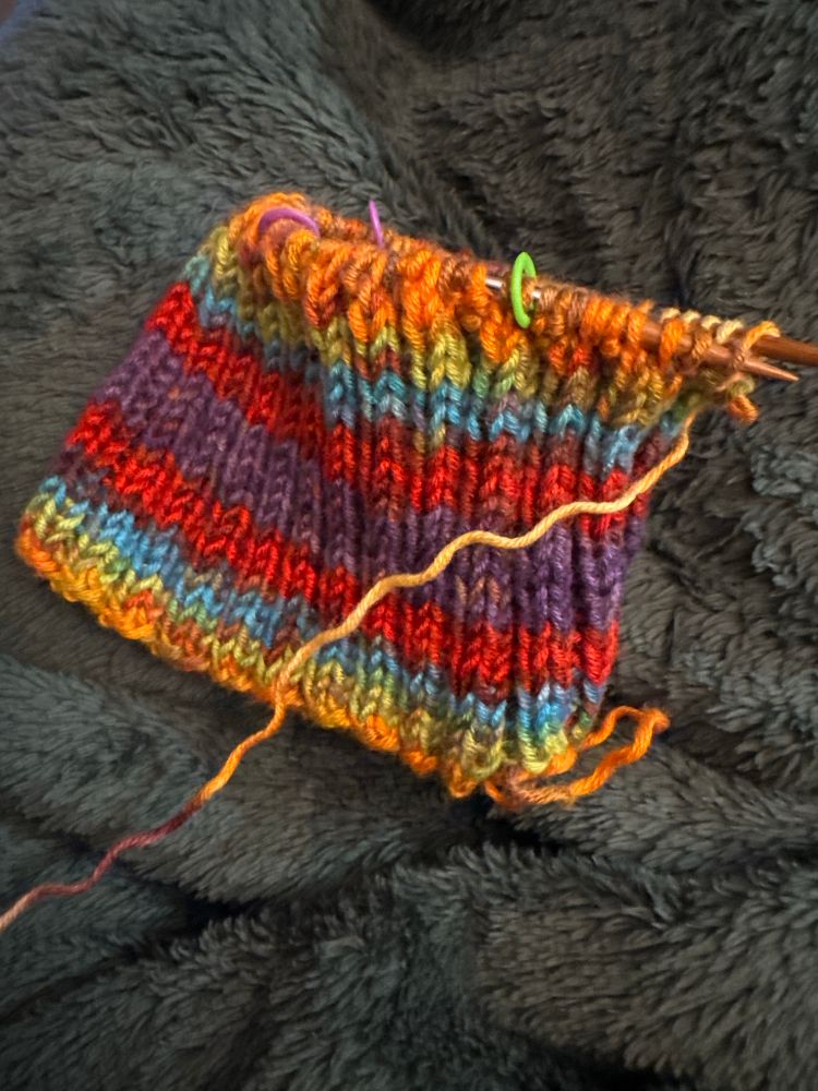 Rainbow colored self-striping yarn in a 2x2 rib pattern forming the bottom rim of a soon-to-be-knitted beanie hat, on very tiny wooden circular needles against a background of a plush dark green blanket. Purple and lime green stitch markers are visible. 