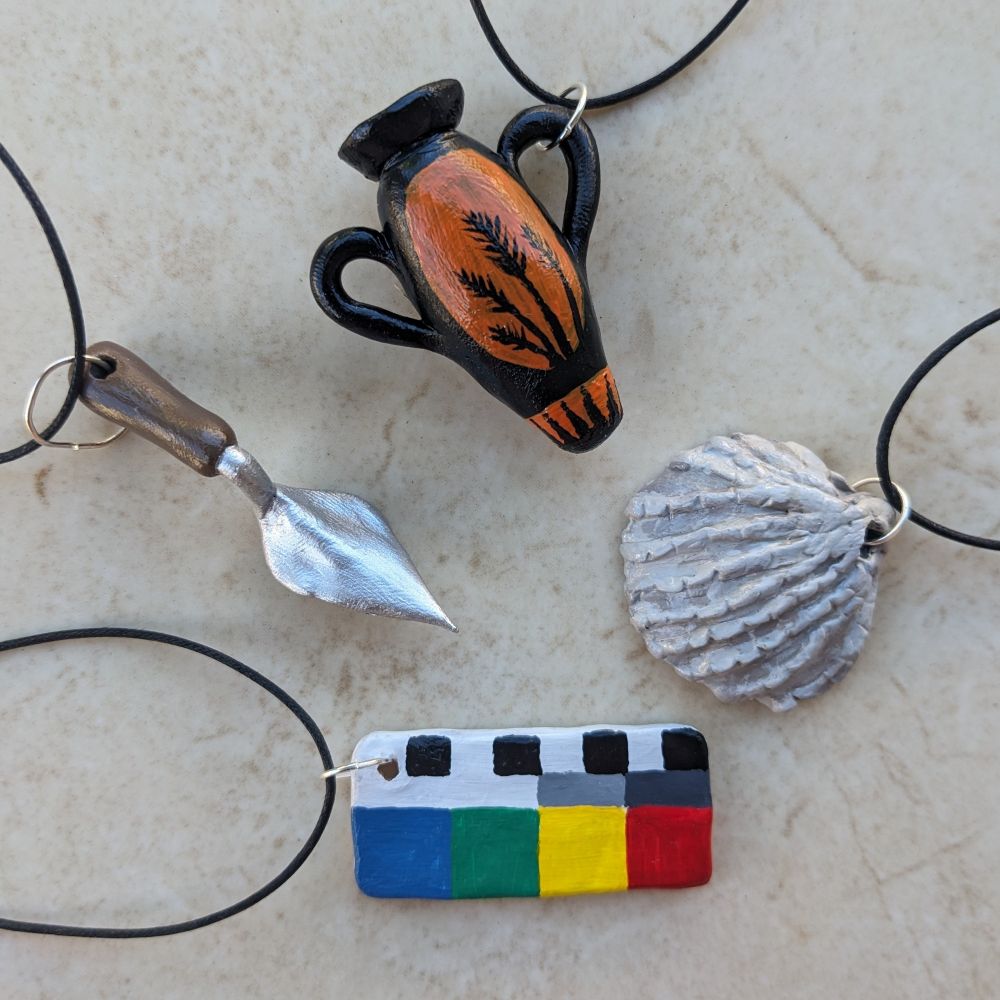 A photograph of four clay pendants of a trowel, an amphora, a bivalve shell half, and a colour scale bar, each attached to loops of black cord