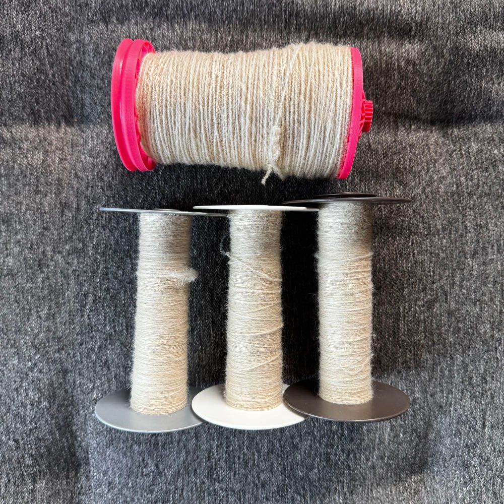 At the top is one bobbin of 2-ply yarn, with 3 storage bobbins with singles below it. The yarn is a blend of white Corriedale, baby camel, and muga silk. The camel and silk add a slight golden sheen