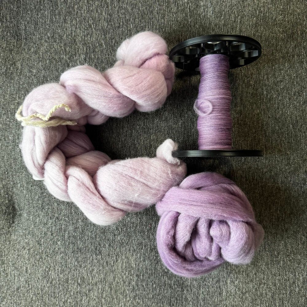 A handspinning project in progress. On the left is a 2 ounce braid of Rambouillet naturally dyed a light lavender. On the right is a 3D printed espinner bobbin (for a Daedalus Starling) with about an ounce of darker purple singles. Below the bobbin is a ball of the remaining darker purple fiber (also naturally dyed on Rambouillet).