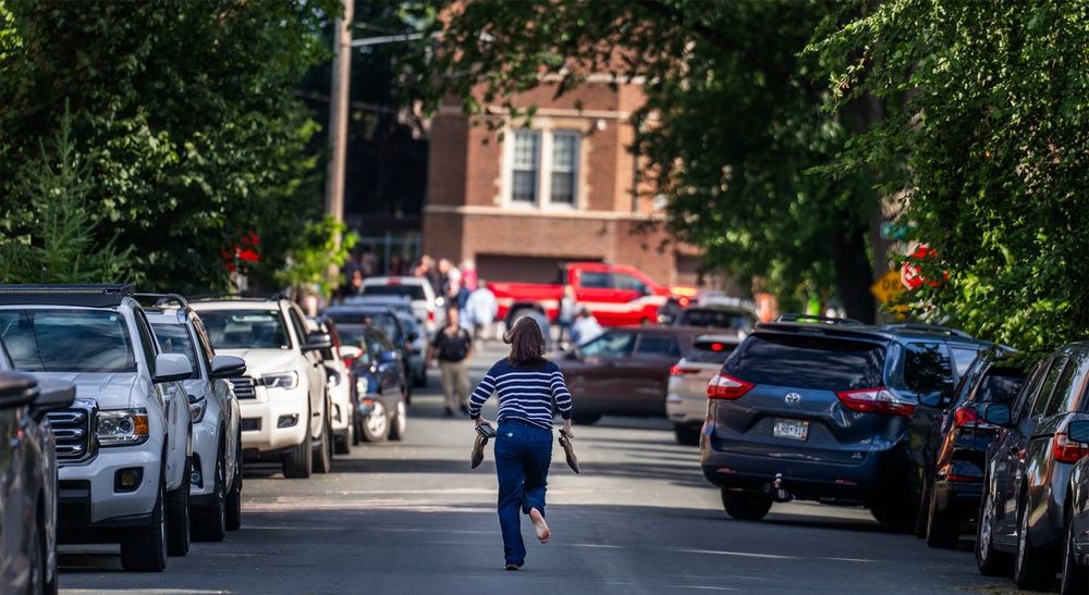An image from the aftermath of the shooting at Annunciation Church in Minneapolis. In the foreground is a woman running in the middle is a street. She is running away from the camera, towards a school in the background. She has her wallet in her hands, and also her shoes, which she presumably took off so she could run faster. You can see the bottom of one of her feet, and dirt is visible (again, presumably from running barefoot).