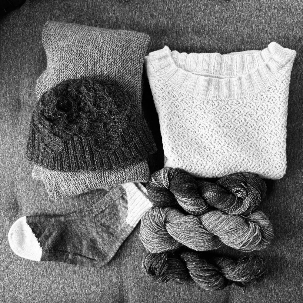 A black and white photo of various handknit projects and handspun skeins of yarn. The projects that appear most distinctive are a white tee with textured lace and a pair of socks with unusual construction and white toes and cuffs.