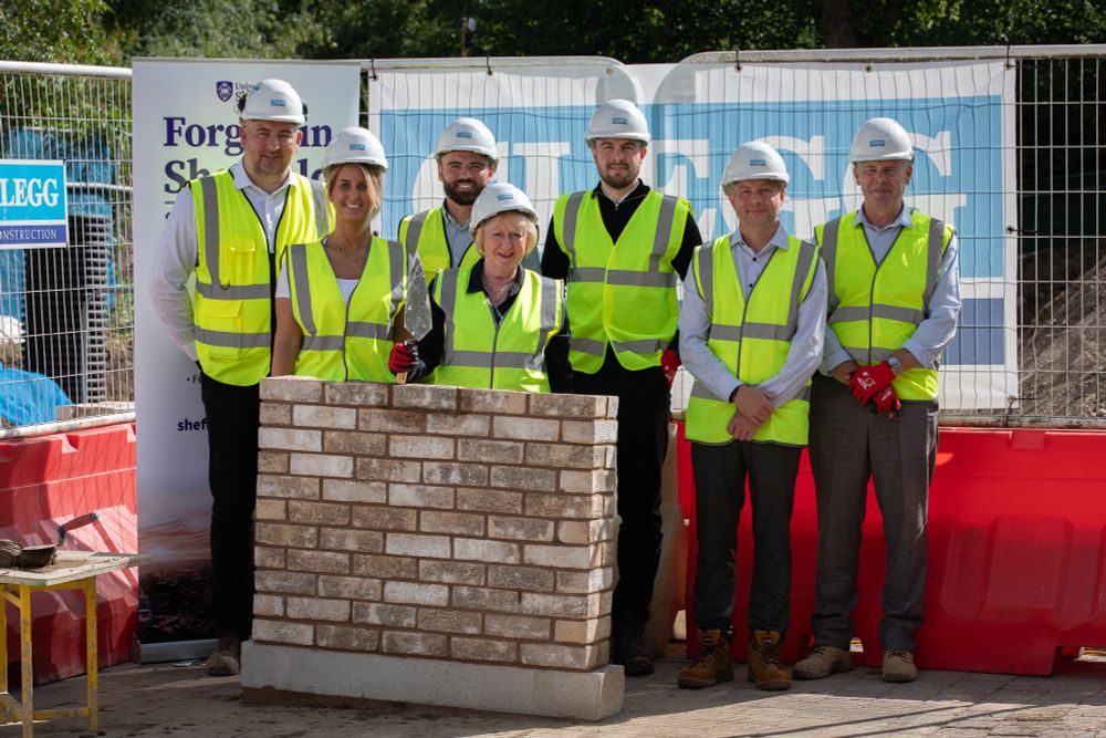 Seven people in high vis vests and hard hats stand behind a section of a wall