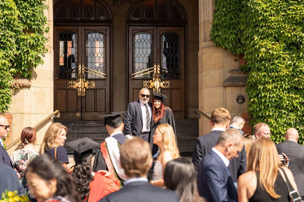 A family member or loved one poses with a graduate in a cap and gown in front of the entrance to Firth Court, with people in the foreground of the shot