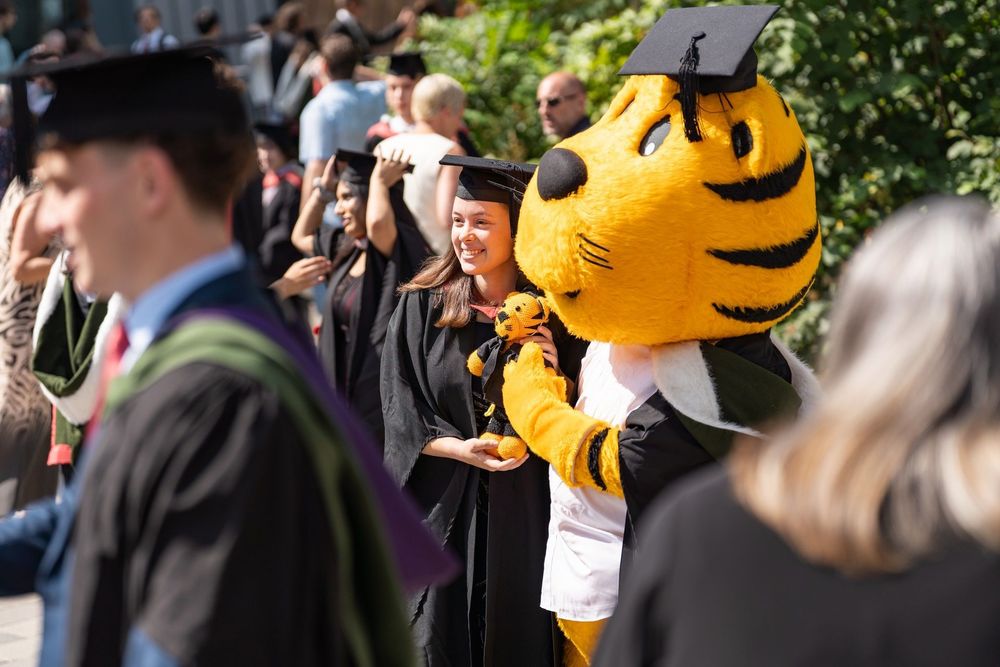 A person in a cap and gown poses with the university mascot, Roary the tiger, whilst holding up a crochet version of Roary the tiger
