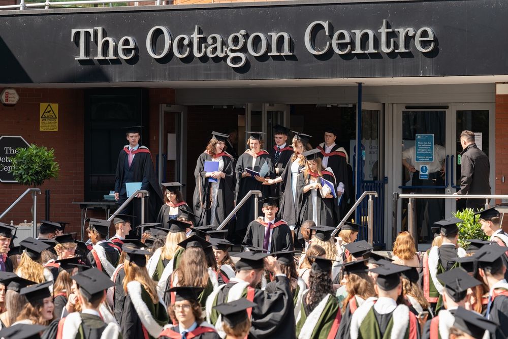 The Octagon Centre entrance, graduates in caps and gowns are walking out the door and joining a crowd of other graduates in caps and gowns

