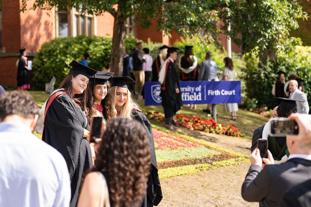 People take photos of three people in graduation caps and gowns, in the background is the purple Firth Court sign, with other graduates surrounding it