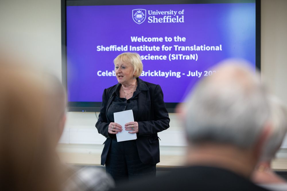 Prof Pamela Shaw stands in front of an audience giving a speech, she wears an all black suit and holds a piece of paper