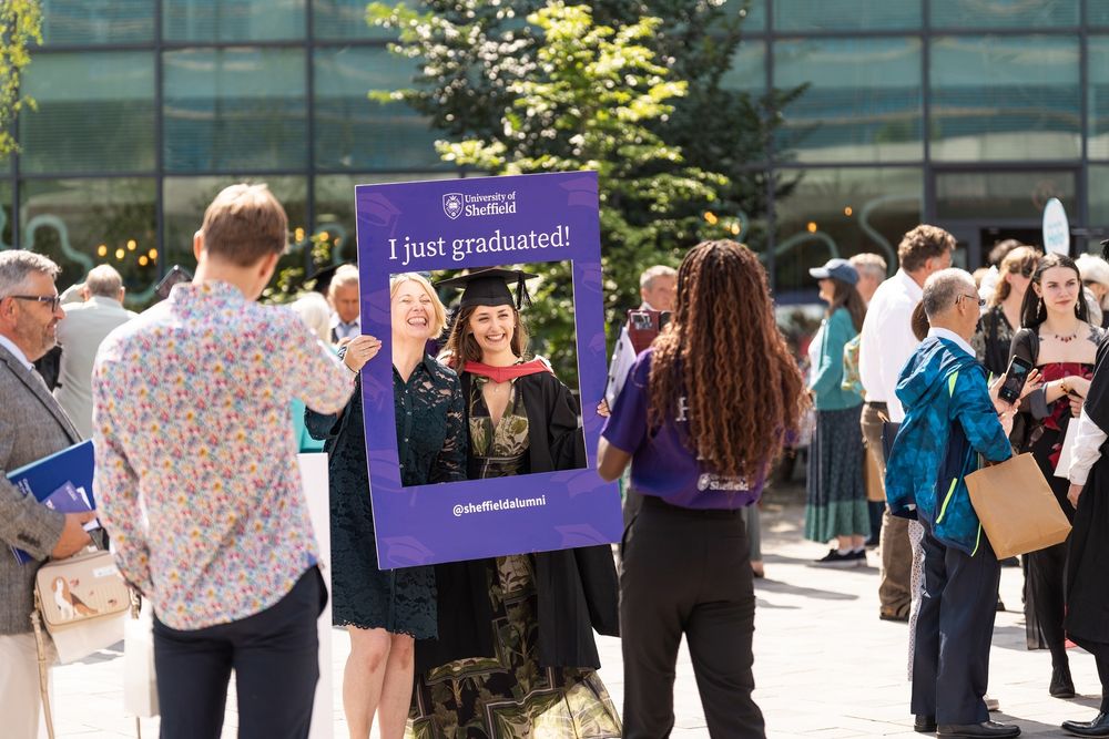 Two people, one in a cap and gown, hold up a purple selfie frame with the words ‘I just graduated’, another person takes their photo