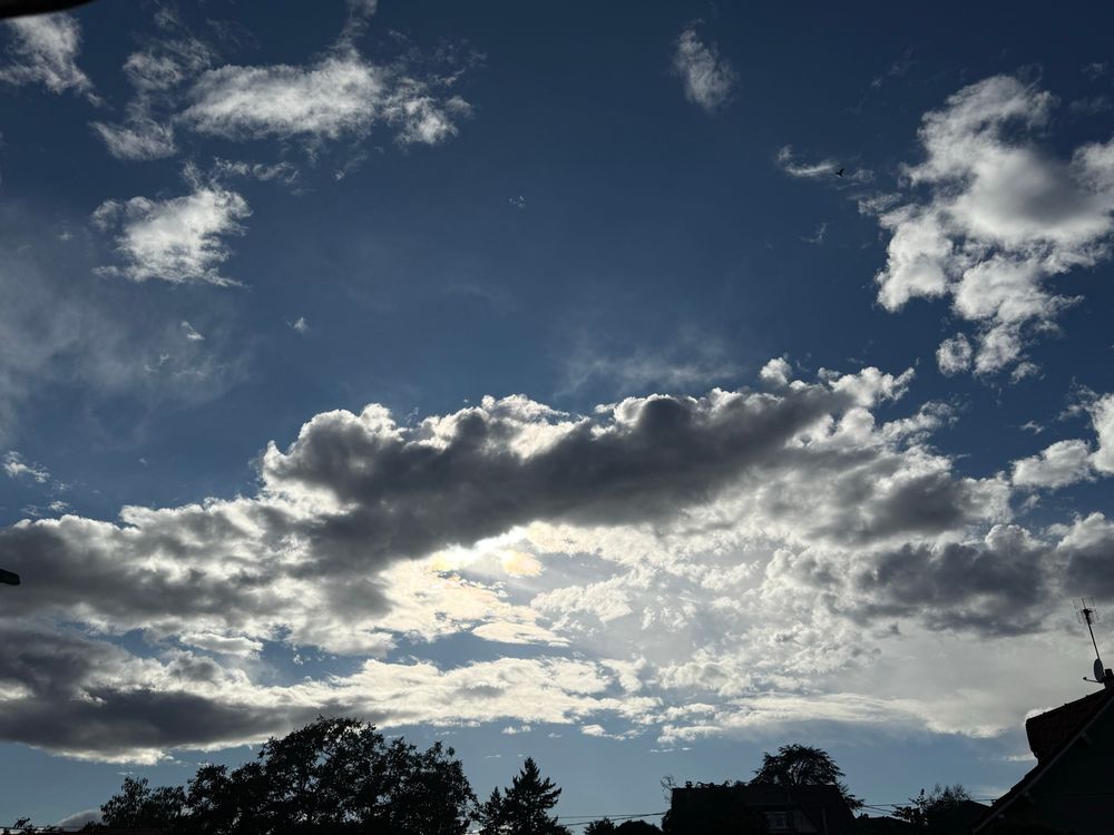 Sur fond de ciel bleu circulent de gros nuages derrière lesquels le soleil joue à cache-cache. Végétations et constructions se dessinent en contrejour. 