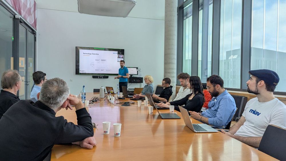 There is a long conference table at the end of the room with people looking at a presentation screen. A man in a blue shirt is presenting to the audience. 