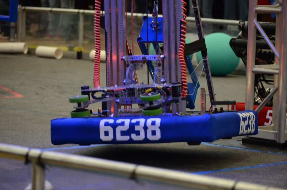 A robot with blue bumpers with the number 6238 printed on them hangs above a carpeted floor. The robot is suspended by a blue metal cage-like structure attached at the back. 