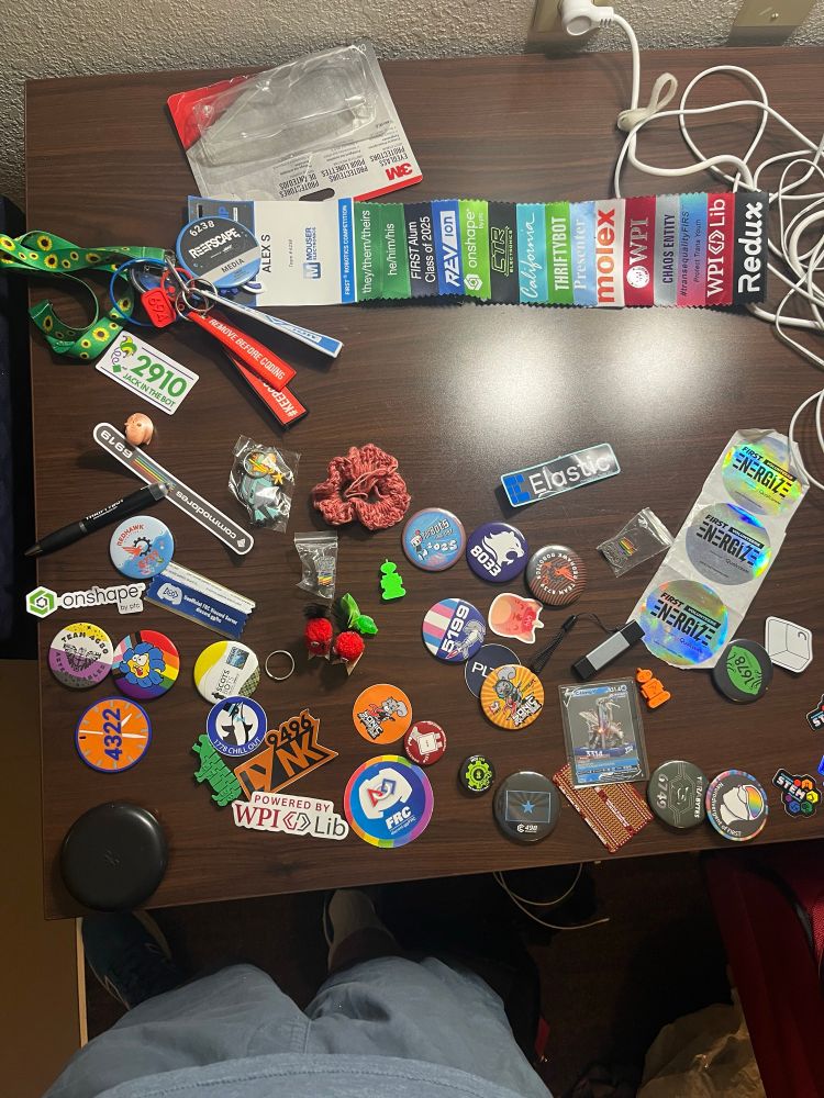Top-down photo of a table with various buttons and stickers scattered on it. At the top of the frame is a name badge with assorted other accoutrements attached to it, including about 10 ribbons. 