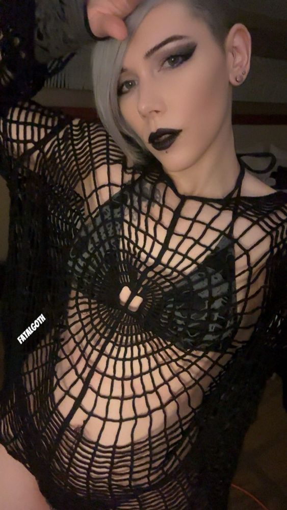 A femme-presenting person with pale skin and silver hair styled in a side shave poses confidently in a black spiderweb-patterned mesh top over a black bikini or lingerie set. They have dramatic black eye makeup, black lipstick, and a piercing below their bottom lip. One arm is raised above their head in a sultry pose. The lighting is dim and moody, enhancing the gothic aesthetic. The username “FATALGOTH” is written on the lower left side.