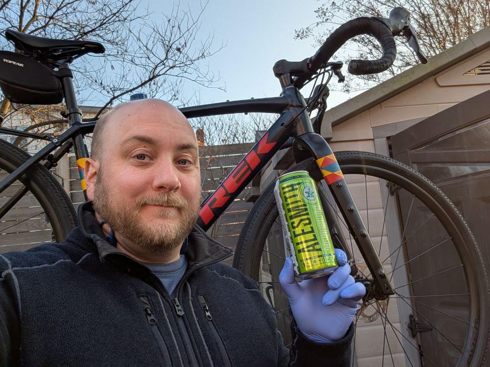 A man looks content holding up a green can of beer in front of a dark blue bicycle that's up in a maintenance stand