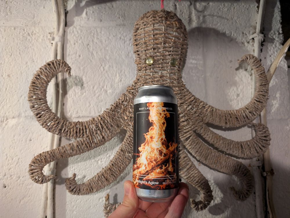A can of beer with a big campfire on the label held up in front of a wicker octopus with circular glass beads for eyes hung on a white cinder block wall