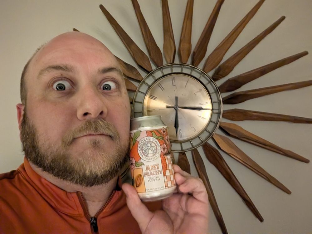 A man in a red bicycle jersey stands in front of a large wooden spoked mid century modern clock with wide crazy eyes with a can of Crooked Stave Just Peachy Fruited Sour Ale in his hand