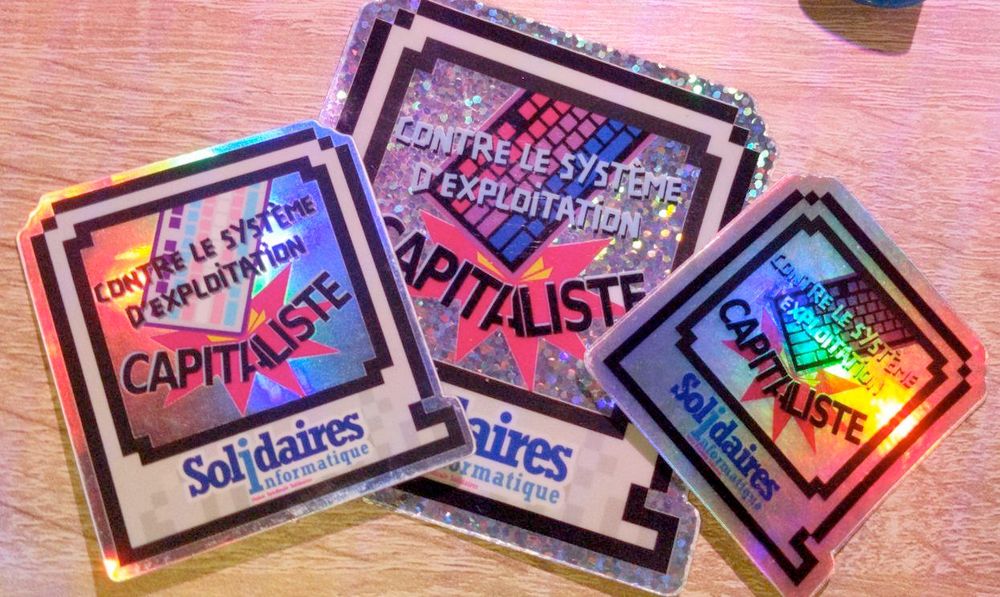 Photo de 3 stickers brillants de taille différentes avec la même forme.

Il est écrit "Contre le système d'exploitation capitaliste" avec un clavier qui casse le mot capitaliste.

L'un des sticker a un clavier aux couleurs du drapeau trans et un autre aux couleurs du drapeau bi.