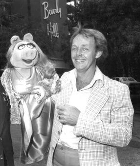 John Lovelady, wearing a white shirt and checkered jacket, smiles as he holds up Miss Piggy in front of a sign that says "Beverly Hills." (Image courtesy of Muppet Wiki)