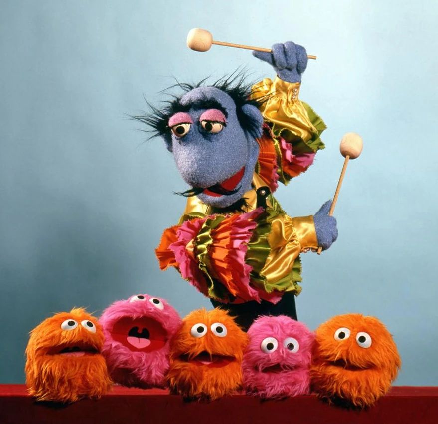 Marvin Suggs, wearing his trademark orange, pink, green, and yellow top, prepares to strike the Muppaphone in front of a pale blue background. (Image courtesy of Muppet Wiki)