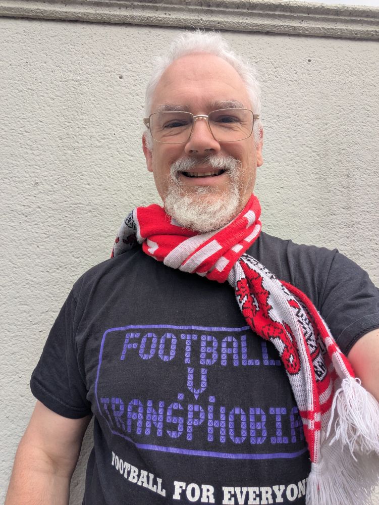 Man with white hair and a beard (me) wearing a red-and-white football scarf with a wyvern on it. The scarf is just about identifiable as a Leyton Orient scarf to those who know. He is wearing a black T-shirt with the words "Football v transphobia: football for all" on it. He is smiling. Is his smile a little apprehensive? He's an Orient supporter; of course it is.