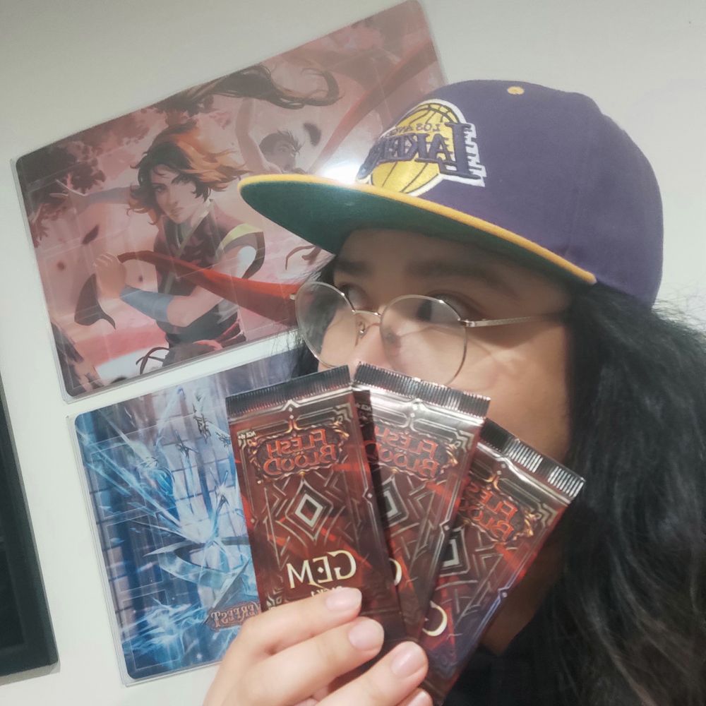 Kiki is holding up three GEM packs courtesy of LSS.
They're wearing a Lakers hat whilst holding the packs in front of their face.

The scarf for a scarf promos mat is visible in the background 