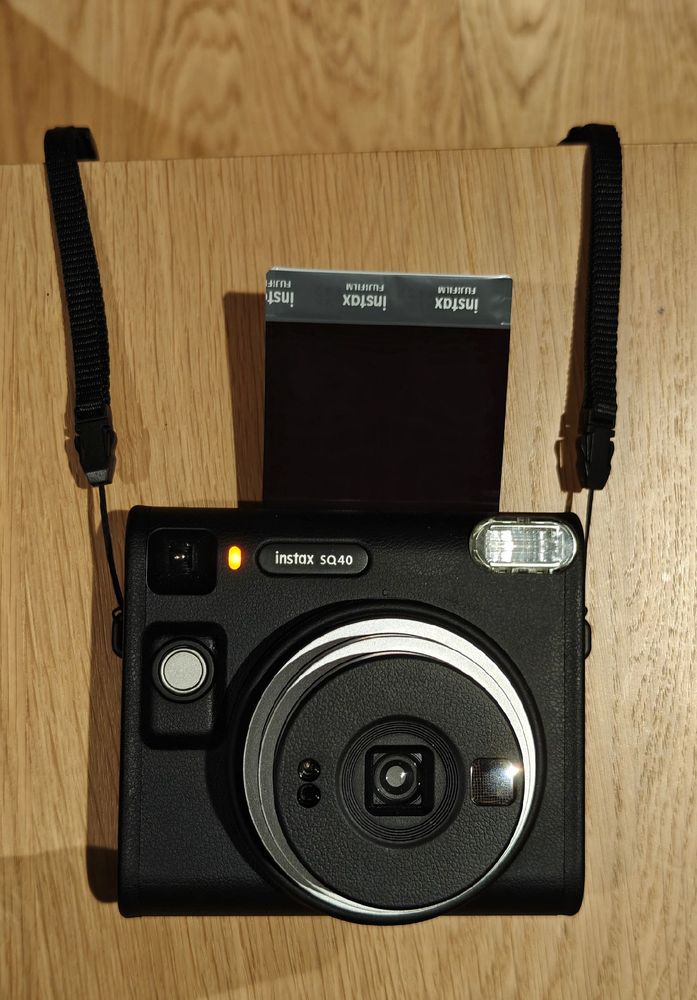 A photo of a black Instax instant camera with a partially ejected film sheet and with wrist straps attached on both sides.