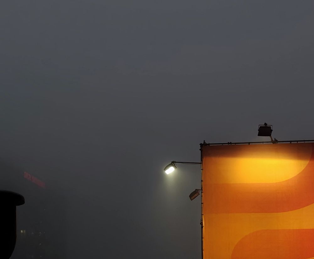 Photo of a large orange wall or billboard illuminated by several mounted lights on a dark, foggy day.