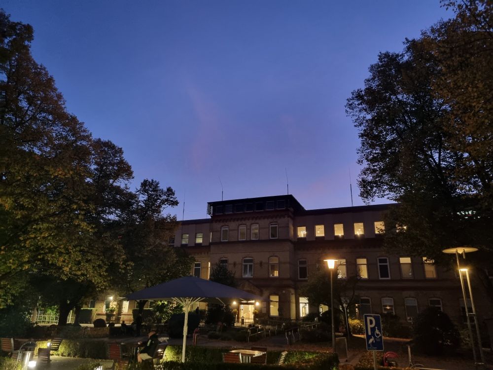 Ein erleuchtetes Gebäude, umrahmt von hohen Bäumen. Abendhimmel in zartviolett im Hintergrund 