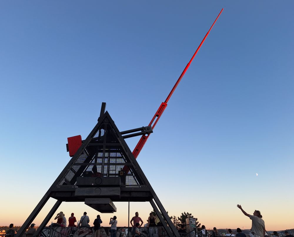 Prague metronome at dusk from behind. Various people below looking at view. One man below half moon gesticulating