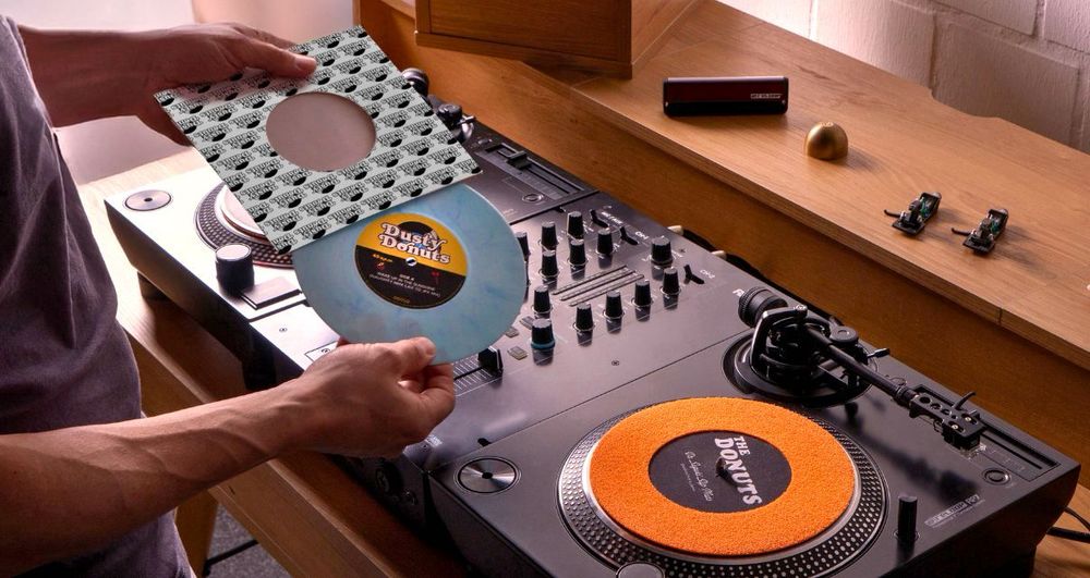 Bild mit zwei 7 inch Plattenspielern und einen Mixer. Eine Hand zieht eine kleine Schallplatte aus ihrer Hülle.