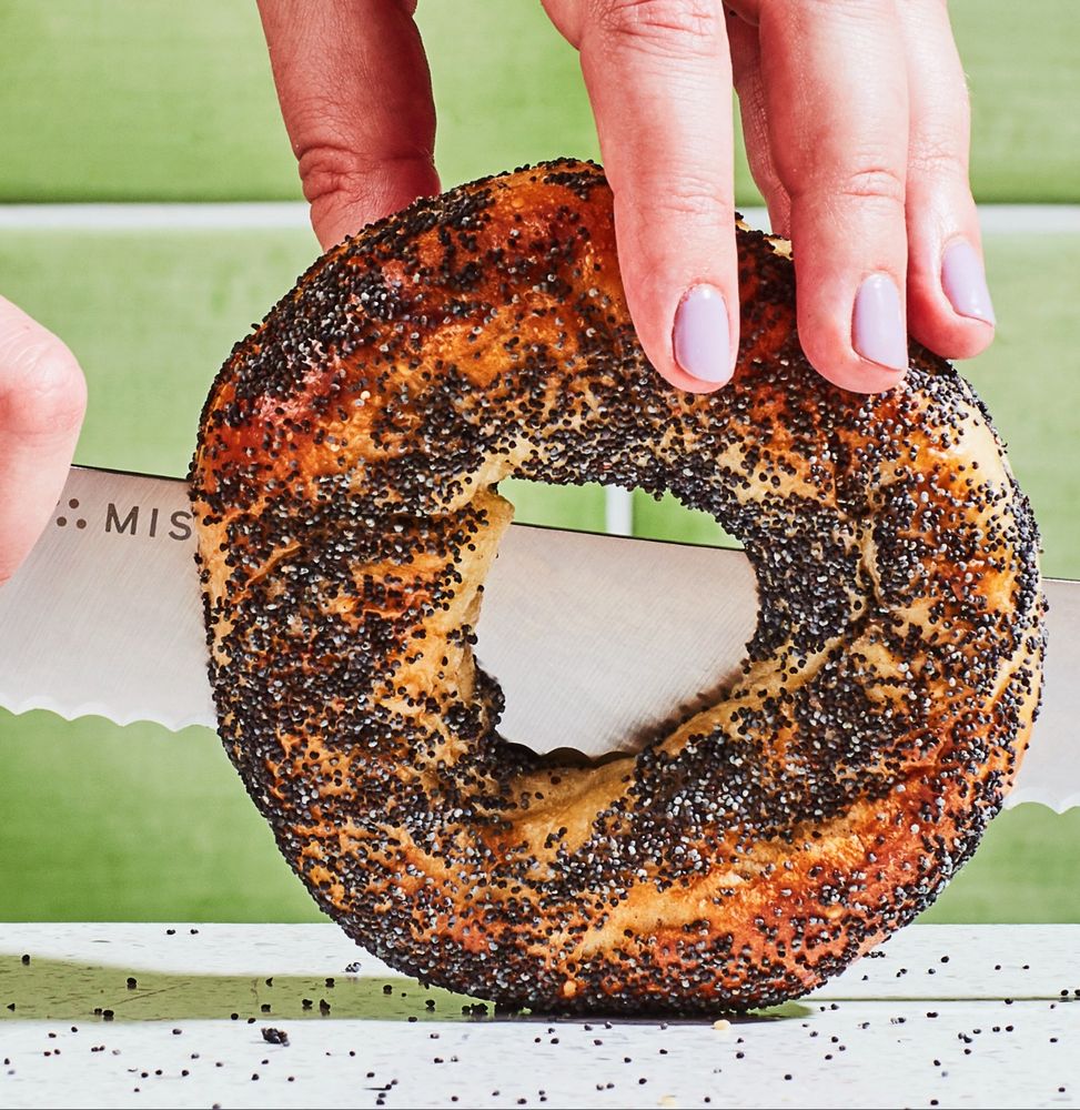 A poppy bagel with a blown out hole being cut by a serrated knife 