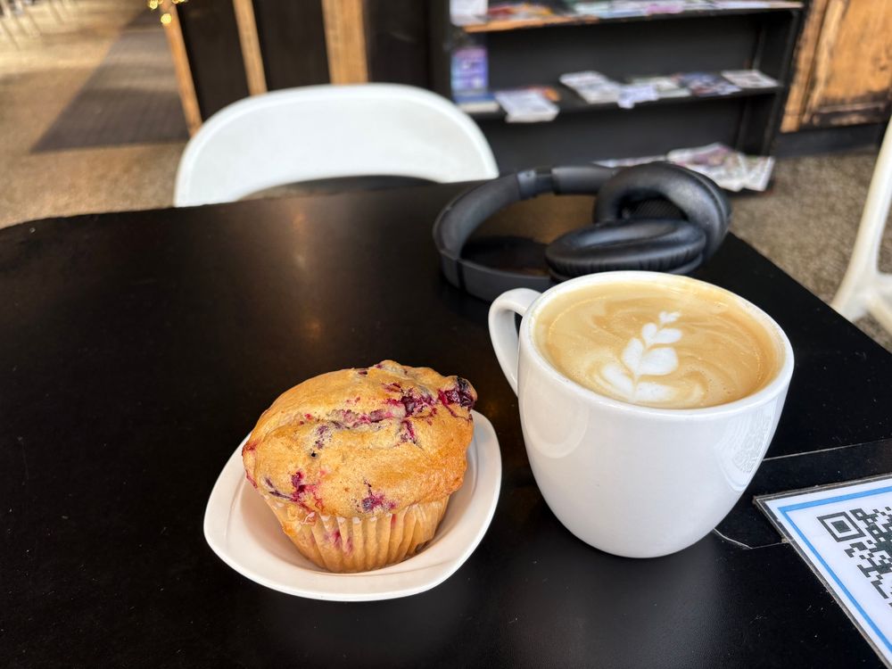 An orange cranberry muffin in a white ceramic dish next to a latte in a white mug on a black table.