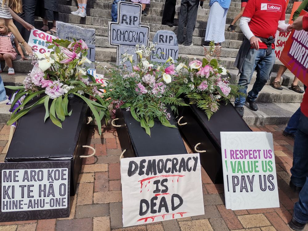 Three black coffins at the bottom of some red brick city steps, with headstones for Trust, Democracy, and Equity. There are flowers on top of the coffins and protest signs surrounding them. 