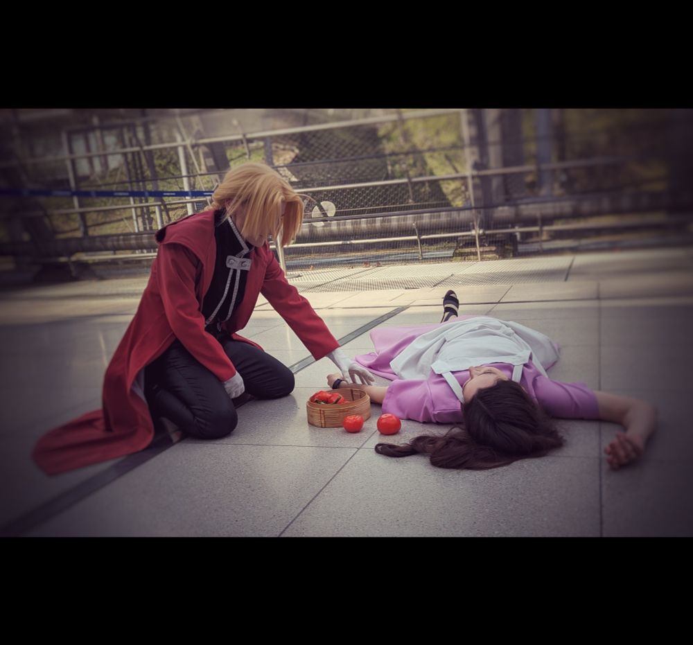 A cosplay photo showing Edward Elric kneeling on the floor in his black clothes, red coat and white gloves. Sprawled in front of him is Trisha Elric in her purple dress and white apron, between them a basket with tomatoes, two having spilled out. Ed is leaking forward slightly, his left hand reaching out, almost touching Trisha's arm, his face is concealed by his bangs.