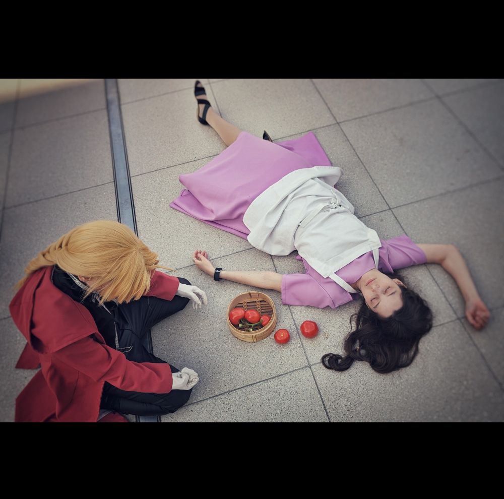 A cosplay photo showing Edward Elric kneeling on the floor in his black clothes, red coat and white gloves. Sprawled in front of him is Trisha Elric in her purple dress and white apron, between them a basket with tomatoes, two having spilled out. Ed's head is lowered, his hair concealing his face. Both his arms are resting atop his thighs, the left hand, close to Trisha's arm, open and limp, the right hand balled into a fist.