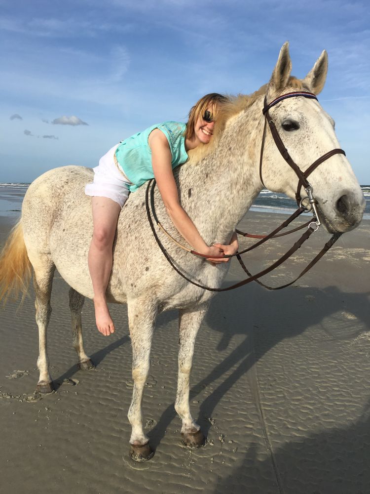 Younger me with medium-length blond hair, a white skirt, and a turquoise shirt, sitting on ATM’s back and leaning forward to hug her neck. ATM, a gray mare wearing a dark brown bridle, looks into the distance as I smile adoringly at her. We are on the wet sand at the beach.