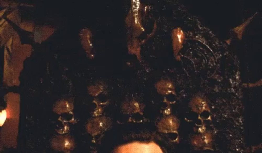 a man in a gold suit is sitting in front of a wall of skulls