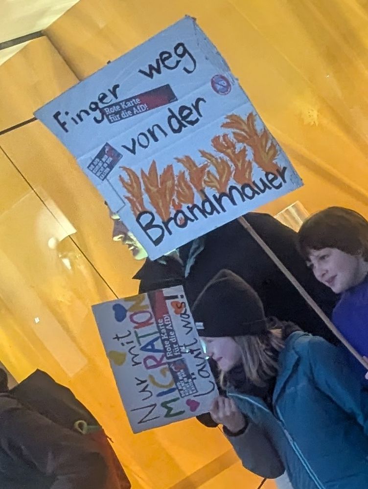 Zwei Demoschilder: 
"Finger weg von der Brandmauer"
und: 
"Nur mit Migration läuft was".
Beide Schilder sind zusätzlich mit zwei Aufklebern beklebt: 
"Rote Karte für die AFD" und "Die AfD kommt mir nicht in den Kasten".