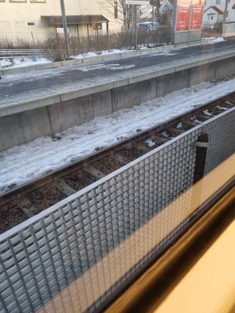 View from a train window. There is a fence in the front of the picture. Behind the fence you can see half a rail track and a platform covered in snow and ice. 