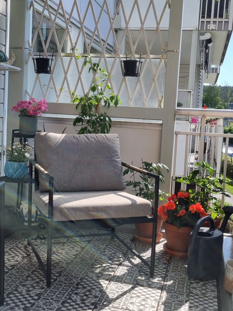 Et bilde av en balkong med blomster, badet i sol. I forgrunnen står en godstolen. Grå og hvite keramikkfliser på gulvet. 