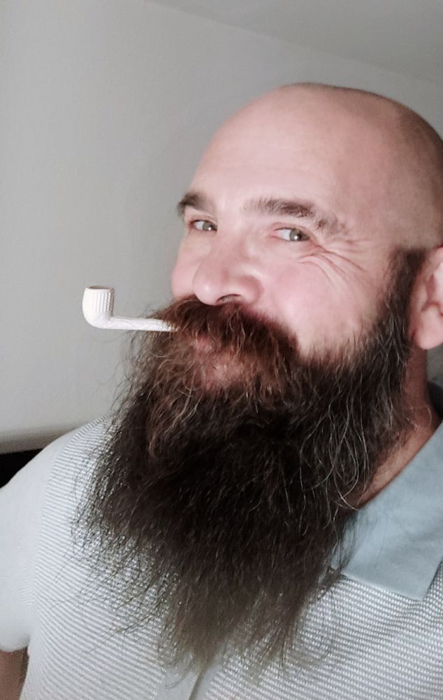 Momakil im Halbprofil in einem hellgrünen Polo. Er grinst in die Kamera und hält eine Tonpfeife von einem Weckmann zwischen den Lippen.