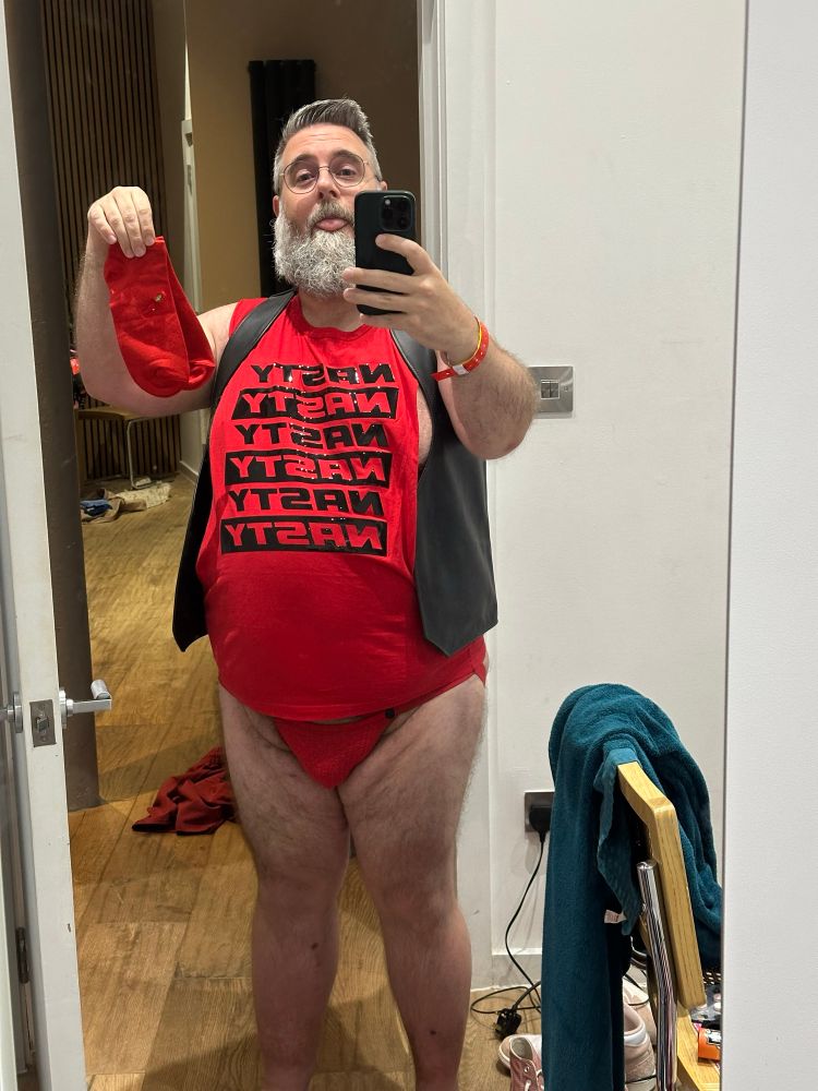 Me wearing a black leather vest over a red tank top with the word “nasty” in large black letters repeated in a single column and a red jockstrap. I’m holding up a pair of red socks with red pikmin on