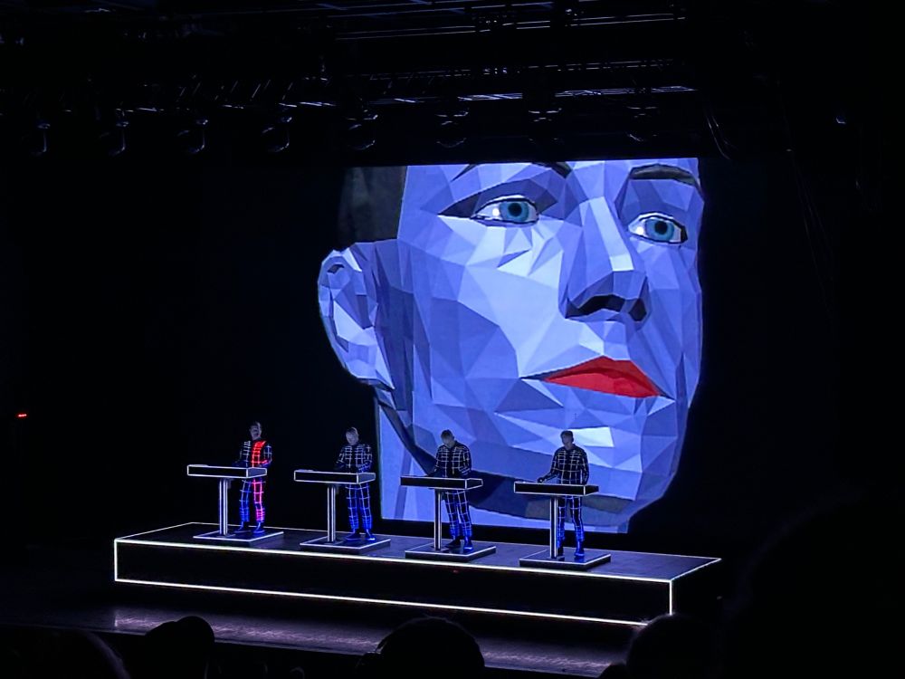 Picture of the Kraftwerk concert in Pittsburgh, March 7, 2025, with the four band members standing at control tables in the front and a big 80s style geometric CGI face on the big screen 