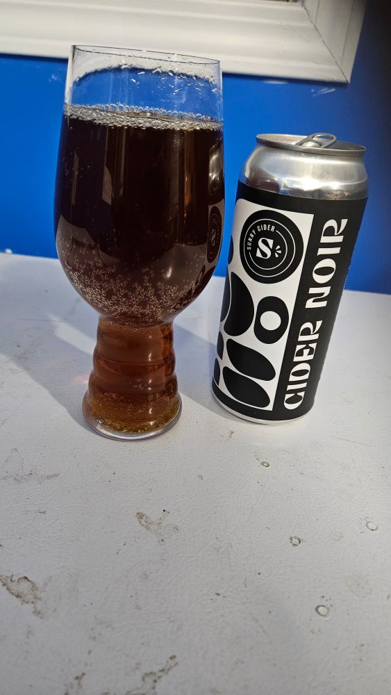 An IPA glass with a brownish liquid, next to a can with the words "Cider Noire" on a white surface and blue background.