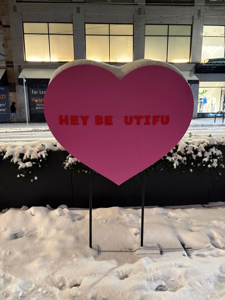 Giant version of a pink candy sweetheart in the snow, with “HEY BE UTIFU”