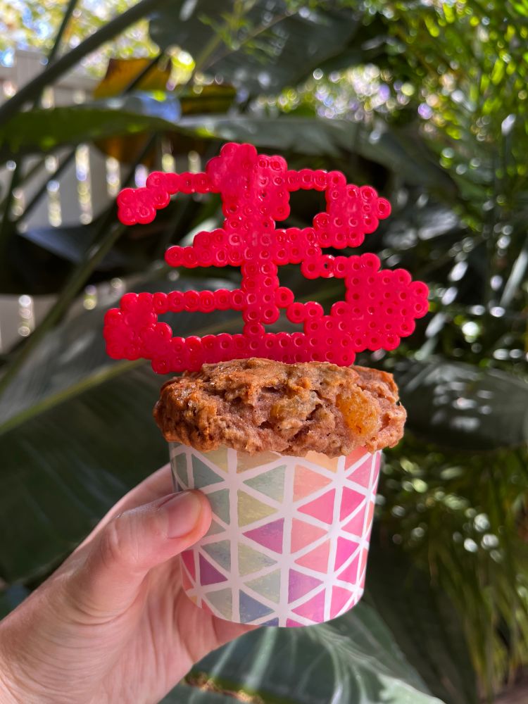 A hand holding a cupcake. The cupcake is decorated with red fuse beads shaped like the letter B in the Buffy font