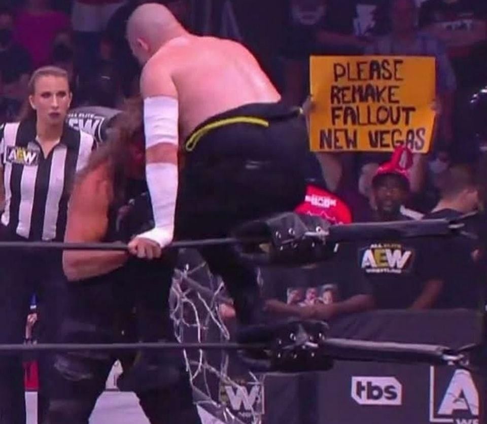 screenshot of wrestling and in the crowd a person is holding a sign that says “please remake fallout new vegas” 
