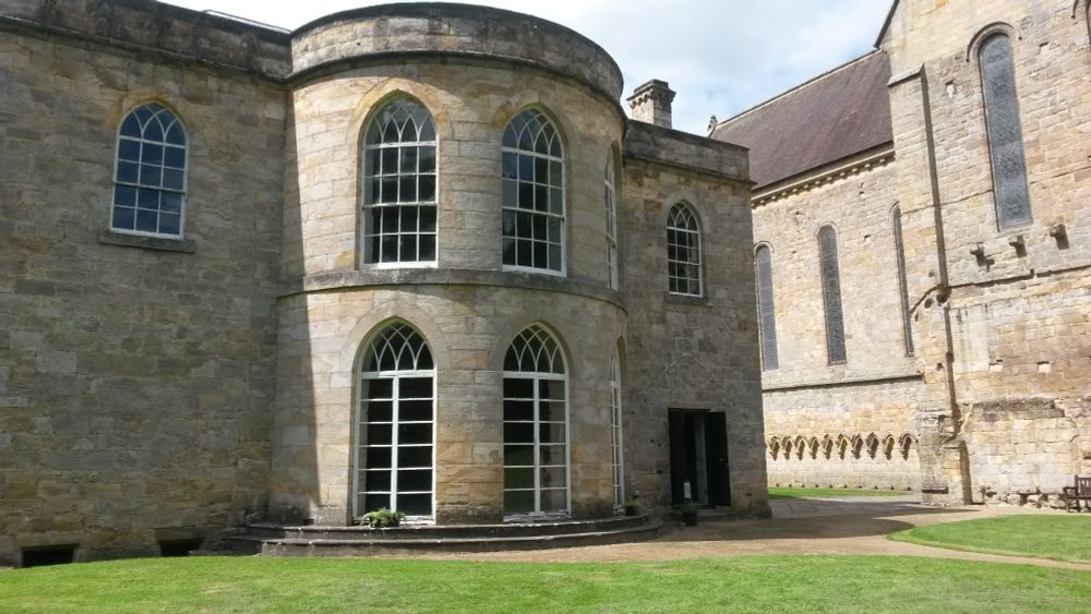 With parts of the south wall and transept of the 12th C Brinkburn Priory church in the background, photo shows eastern extension of manor house built post dissolution of monasteries. 
A semi-circular two storey feature has three full height glazed doors on the ground floor opening onto the lawn, and windows of almost the same size on the floor above. These are all in a Georgian style with intersecting tracery arches at the top. Two further windows are in the wall either side of the curved section, also with pointed arches. There is a further wooden door to the right.